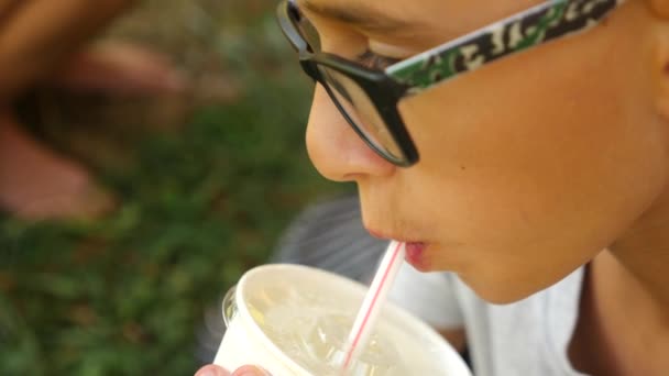 Привлекательный мальчик пьет напиток через соломинку, сидя на траве под деревом в парке. 4К, замедленная съемка — стоковое видео
