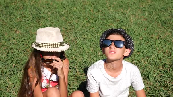 Мальчик и девочка, близнецы 10 - 12 лет сидят на поляне в парке в летний солнечный день. радуйтесь и обнимайтесь. 4k, крупный план, замедленная съемка, копировальное пространство — стоковое видео