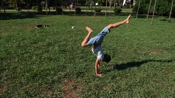 Забавный юный кавказский мальчик танцует на зеленой траве под открытым небом в летний солнечный день. 4k, slow motion — стоковое видео