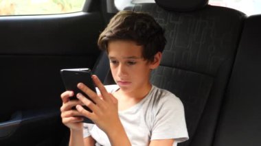 Araba koltuğuna oturan ve arka koltukta yolculuk yaparken akıllı telefonda oynamak güzel çocuk. 4k, ağır çekim