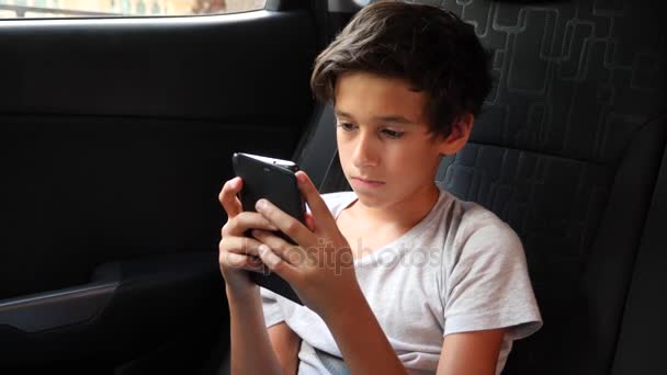 Schöner Junge, der im Autositz sitzt und auf dem Smartphone spielt, während er auf dem Rücksitz mitfährt. 4k, Zeitlupe — Stockvideo