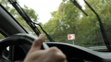 Yağmurda arabada ön cam yoldan görünümünü. Su damlaları görüntülemek zorlaştırır camına düşüyor. 4k, ağır çekim. adam direksiyon tutan.