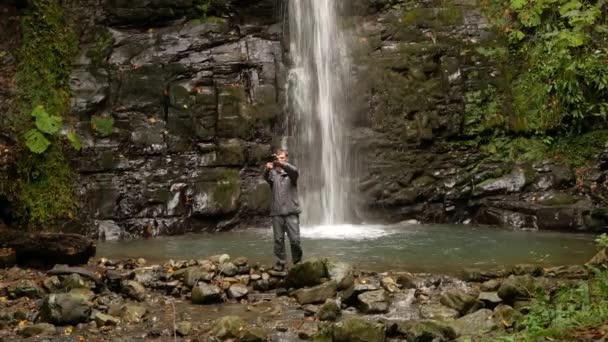 A man is a tourist standing against a waterfall. 4k takes himself to the camera. — Stock Video