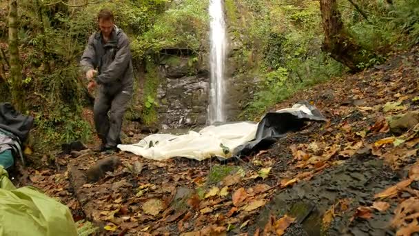 4k. un turista masculino organiza una carpa al pie de la montaña sobre el telón de fondo de una cascada . — Vídeos de Stock