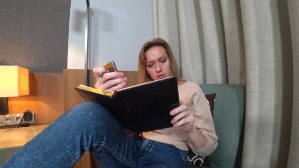 Por la noche, la mujer se sienta en un cómodo sillón en la sala de estar, habla en video desde su teléfono inteligente y toma notas en un cuaderno. 4k — Vídeo de stock