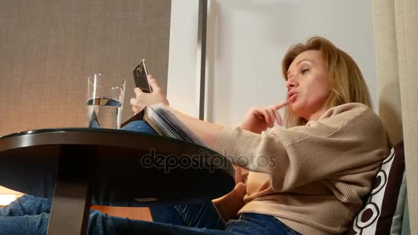 Le soir, la femme s'assoit sur un fauteuil confortable dans le salon, parle sur vidéo depuis son smartphone et prend des notes dans un carnet. 4k — Video