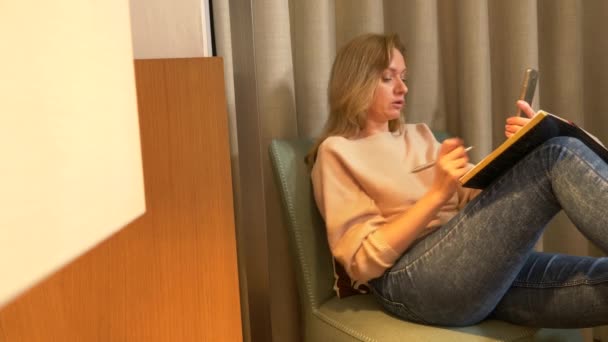 Wieczorem, kobieta siedzi na wygodny fotel w pokoju dziennym, rozmowy na wideo od jej smartfona i sprawia, że notatki w notesie. 4k — Wideo stockowe