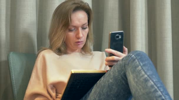 Večer, žena sedí na pohodlné křeslo v obýváku, mluví na video ze svého smartphone a napíše do poznámkového bloku. 4k — Stock video