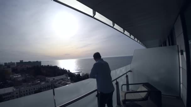 Ο άνθρωπος στέκεται στο μπαλκόνι για ένα ηλιόλουστο πρωί και κοιτάζει προς τα κάτω για την πόλη και την παραλία. 4k — Αρχείο Βίντεο
