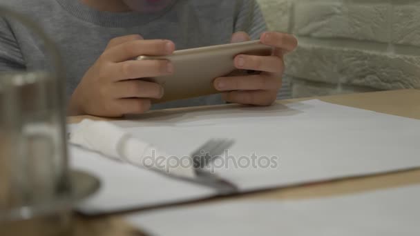 Un niño está jugando con un teléfono móvil en la mesa, esperando un pedido en un café. niño sentado en la mesa, sosteniendo el teléfono móvil moderno, y presionando su dedo en la pantalla táctil. Primer plano, 4k — Vídeos de Stock