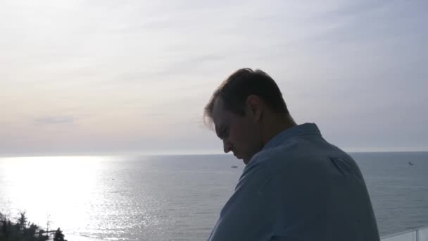 L'uomo è in piedi sul balcone in una mattina di sole e guarda giù sulla città e la riva del mare. 4k — Video Stock