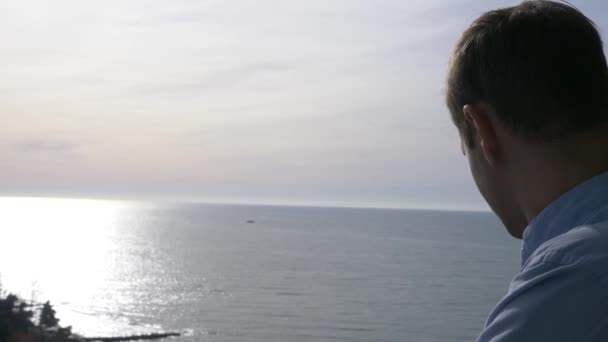 Der Mann steht an einem sonnigen Morgen auf dem Balkon und blickt auf die Stadt und das Meer. 4k — Stockvideo