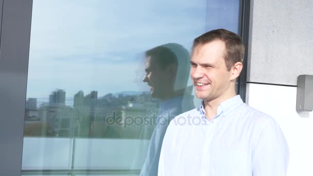 De gelukkige man staat op het balkon in de buurt van het venster waarin de stad met moderne hoogbouw. op een heldere, zonnige dag. 4k — Stockvideo