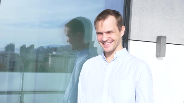 O homem sortudo está em pé na varanda perto da janela, o que reflete a cidade com edifícios modernos de arranha-céus. num dia claro e ensolarado. 4k — Vídeo de Stock