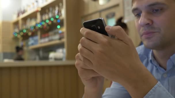 Csinos, fiatal férfi egy okostelefon, kávét iszik, és mosolyogva, miközben egy kávézóban lazíthatnak. Karácsonykor. 4k — Stock videók