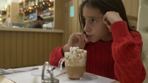 Une fille brune avec des boissons aux cheveux longs avec plaisir chocolat chaud avec guimauve et crème fouettée dans un café. Noël. 4k — Video