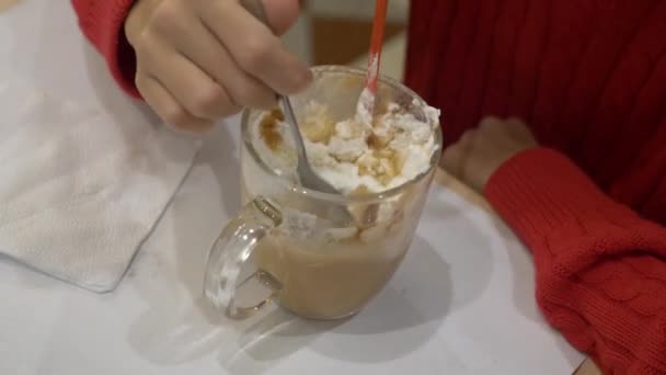 Une fille brune avec des boissons aux cheveux longs avec plaisir chocolat chaud avec guimauve et crème fouettée dans un café. Noël. 4k — Video