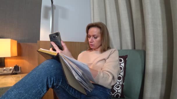 In the evening, the woman sits on a comfortable armchair in the living room, talks on video from her smartphone and makes notes in a notebook. 4k — Stock Video