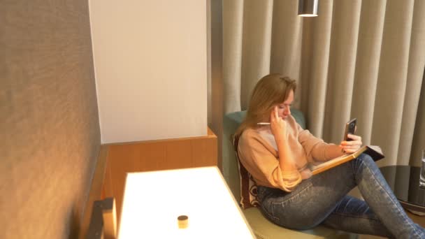 저녁에 여자는 거실에 있는 안락한 안락의 자에 앉아, 그녀의 smartphone에서 비디오에 이야기 고 노트 노트북 합니다. 4 k — 비디오