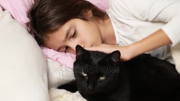 Una niña pequeña abraza y acaricia a su mascota, un gato negro, acostado en la cama. 4k , — Vídeos de Stock