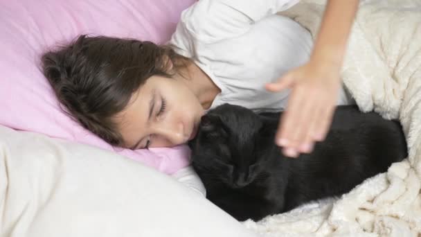 Una niña pequeña abraza y acaricia a su mascota, un gato negro, acostado en la cama. 4k , — Vídeos de Stock