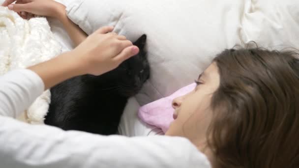 Ein kleines Mädchen umarmt und streichelt ihr Haustier, eine schwarze Katze, die im Bett liegt. 4k, — Stockvideo