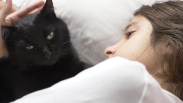 Una niña pequeña abraza y acaricia a su mascota, un gato negro, acostado en la cama. 4k , — Vídeos de Stock