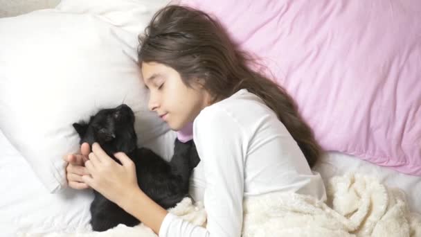 En liten flicka kramar och smeker sin husdjur, en svart katt, liggande i sängen. 4k, — Stockvideo