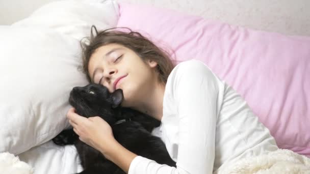 Una niña pequeña abraza y acaricia a su mascota, un gato negro, acostado en la cama. 4k , — Vídeos de Stock