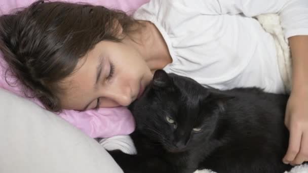 Маленька дівчинка обіймає і гладить свого улюбленця, чорного кота, лежачого в ліжку. 4k , — стокове відео