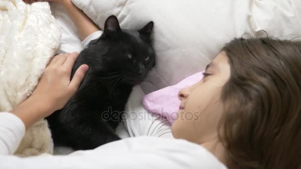Маленька дівчинка обіймає і гладить свого улюбленця, чорного кота, лежачого в ліжку. 4k , — стокове відео