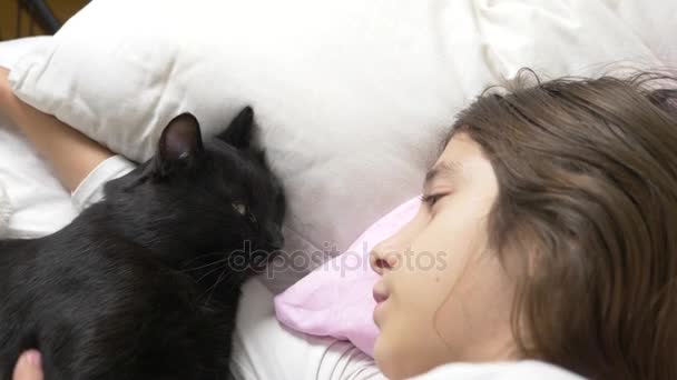 Een klein meisje knuffels en lijnen van haar huisdier, een zwarte kat, liggend in bed. 4k, — Stockvideo