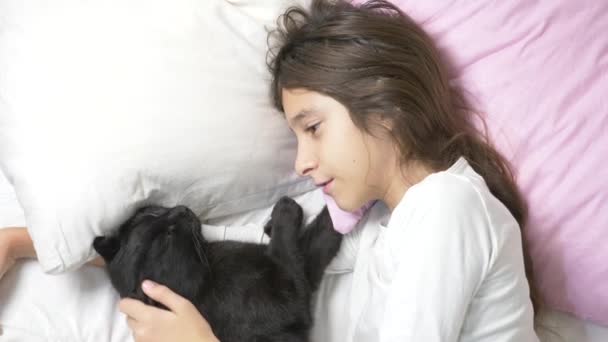 Une petite fille étreint et caresse son animal de compagnie, un chat noir, couché au lit. 4k , — Video