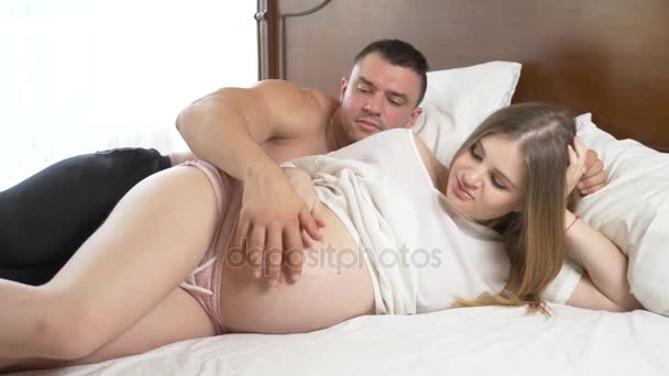 Una hermosa joven embarazada y su musculoso marido yacen en la cama, el marido está planchando el vientre de su esposa. 4k, cámara lenta — Vídeo de stock