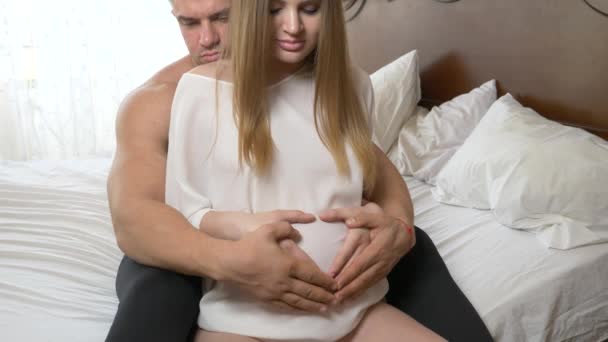 Красивая молодая беременная женщина и ее мускулистый муж лежат на кровати, муж гладит брюхо своей жены. 4k, slow motion — стоковое видео