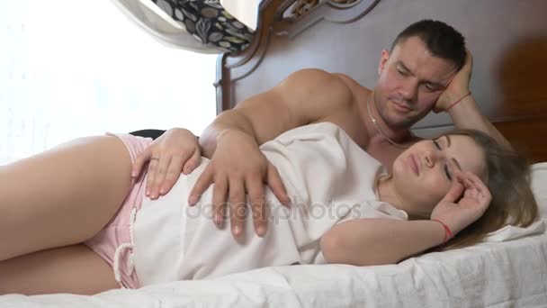 Μια όμορφη νεαρή έγκυος γυναίκα και ο σύζυγός της μυϊκής ψέμα στο κρεβάτι, ο σύζυγος σιδερώματος την κοιλιά της συζύγου του. 4k, αργή κίνηση — Αρχείο Βίντεο