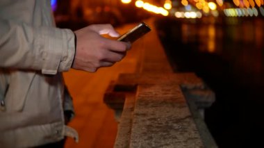 Bir adam Rıhtımlar üzerinde duruyor ve gece gece ışıkları çerçevede bir telefon kullanır. 4k, arka plan bulanıklık