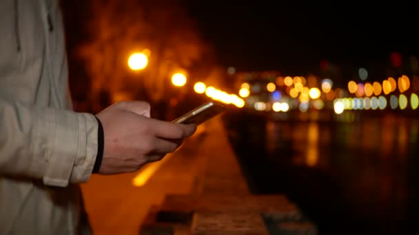 Un uomo si trova sul lungomare e usa il telefono di notte su uno sfondo di luci notturne. 4k, sfocatura dello sfondo — Video Stock