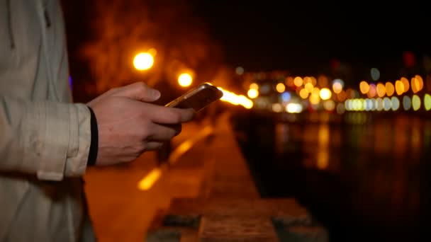 Um homem fica à beira-mar e usa o telefone à noite contra um fundo de luzes noturnas. 4k, borrão de fundo — Vídeo de Stock