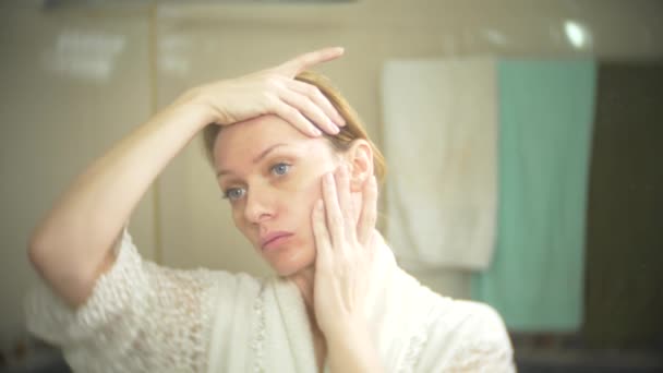 Middelbare leeftijd vrouw die haar huid in een spiegel kijkt. vrouw zoekt rimpels op haar gezicht. 4k, slow-motion — Stockvideo