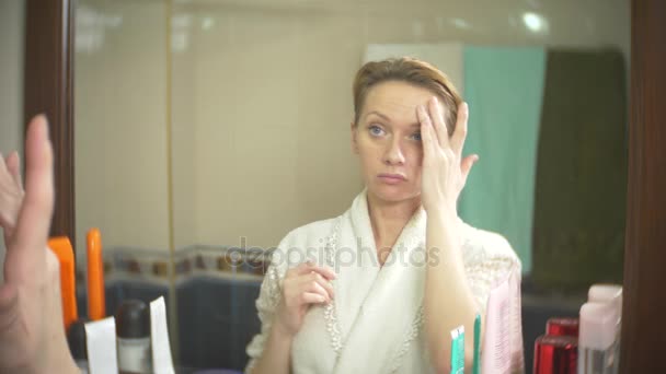 Femme d'âge moyen regardant sa peau devant un miroir. femme regarde les rides sur son visage. 4k, au ralenti — Video