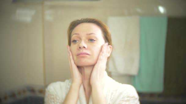 Женщина средних лет смотрит на свою кожу перед зеркалом. У женщины морщины на лице. 4k, slow motion — стоковое видео