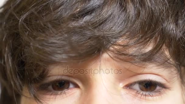 En pojke med långa svarta ögonfransar bruna ögon. Han ser ut från under en lång lockigt luggen. 4 k, Slowmotion, närbild — Stockvideo