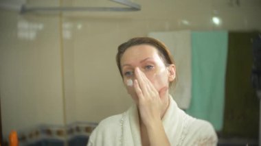 yüz ve boyun banyoda krem uygulayarak bornoz kadında. 4k, ağır çekim