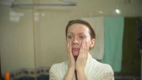 Γυναίκα στο μπουρνούζι εφαρμογή κρέμας σε πρόσωπο και λαιμό στο μπάνιο. 4k, αργή κίνηση — Αρχείο Βίντεο