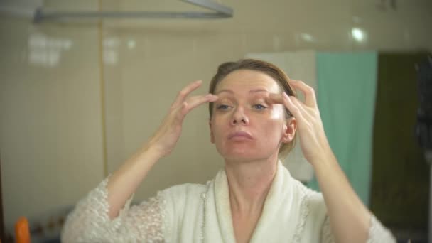 Frau im Bademantel cremt Gesicht und Hals im Badezimmer ein. 4k, Zeitlupe — Stockvideo