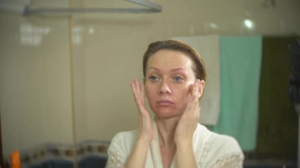 Woman in bathrobe applying cream on face and neck in bathroom. 4k, slow motion — Stock Video