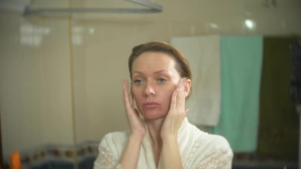 Mujer en albornoz aplicar crema en la cara y el cuello en el baño. 4k, cámara lenta — Vídeos de Stock