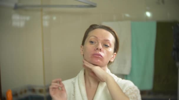Frau im Bademantel cremt Gesicht und Hals im Badezimmer ein. 4k, Zeitlupe — Stockvideo