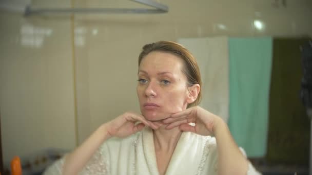 Femme en peignoir appliquer la crème sur le visage et le cou dans la salle de bain. 4k, au ralenti — Video